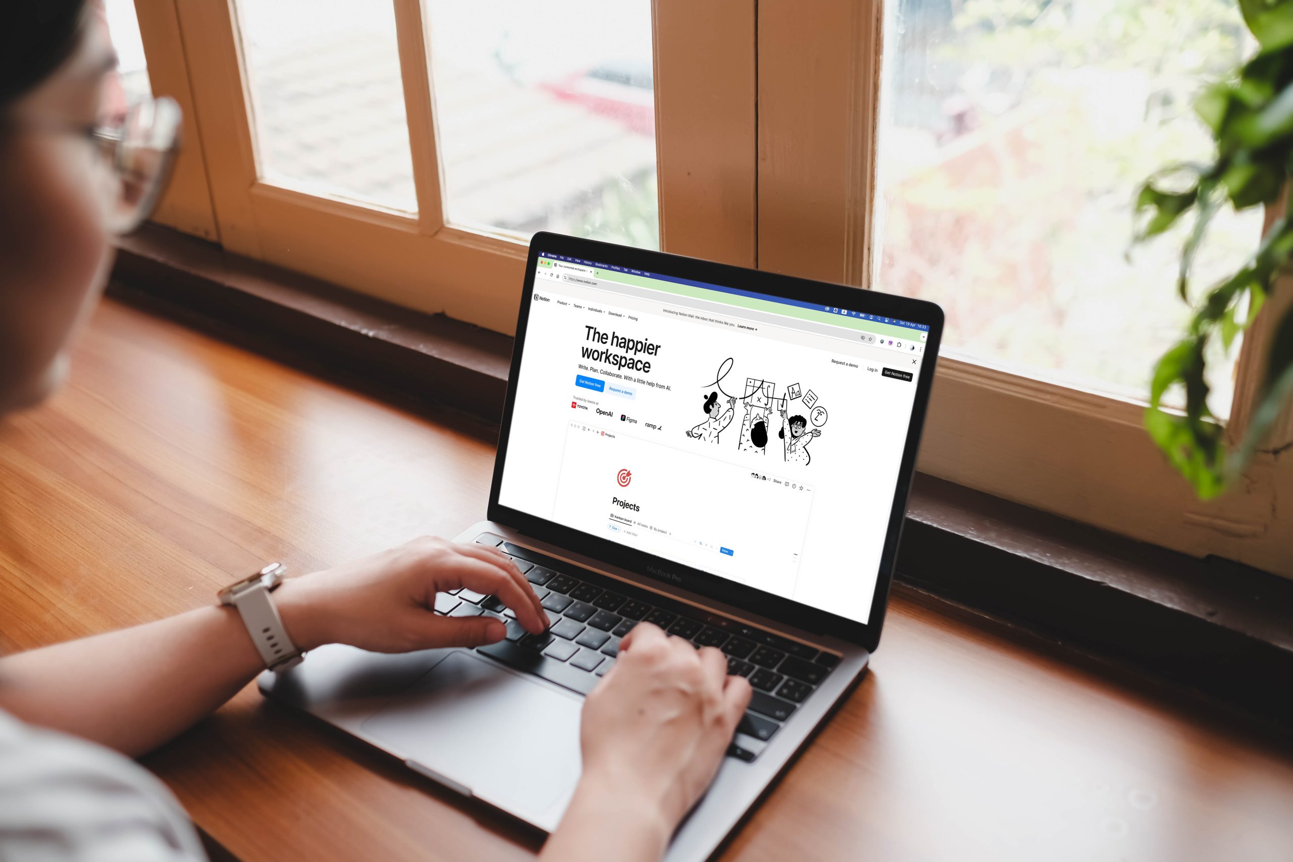 A woman organizing her projects on the Notion productivity app on a laptop, working at a wooden desk by a window.