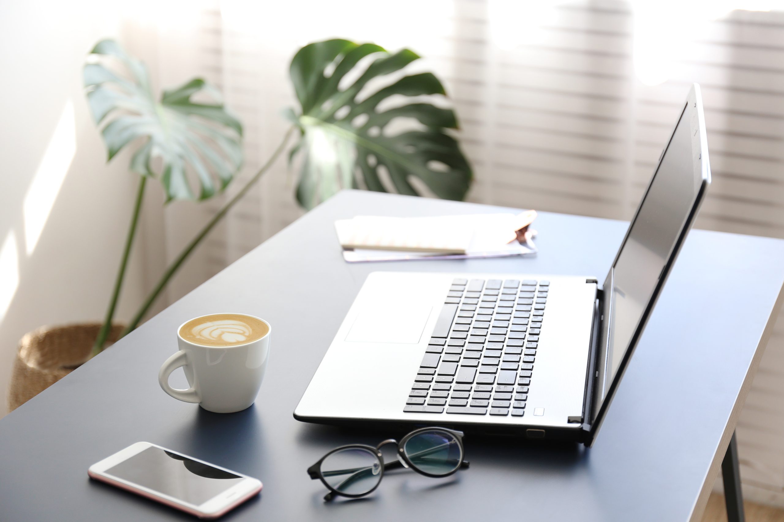 Feminine office space with a lot of ntural light. Office interior design concept. White laptopp with black keyboard, desk w/ matte blue table top. Computer, blank skreen smartphone. Freelance blogger writing an article. Close up, copy space for text.